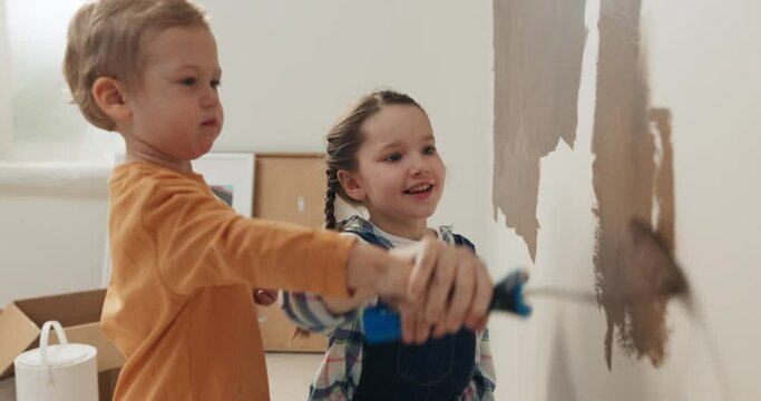 Seven-year-old girl with and two-year-old boy with blond hair are holding rollers and painting wall in brown. Children are smiling. Boy is wearing orange sweater, and girl is wearing checkered shirt.