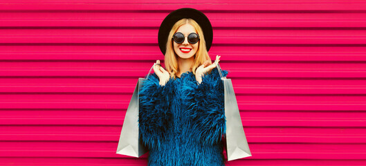 Portrait of beautiful stylish smiling woman with shopping bags wearing blue fur coat, black round hat and sunglasses posing on pink background