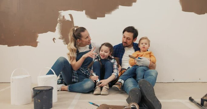 Man, wife and two children are sitting on floor in new house. The family paints walls in brown. They are dressed in jeans and checkered shirts, chatting and smiling. Boy and girl hold rollers in hands