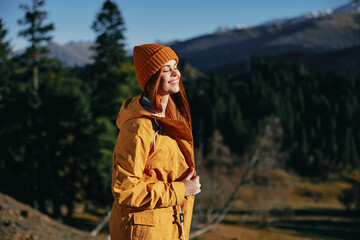 Woman full-length hiker smiling standing with her back to the mountain looking at nature happiness overlooking snowy mountains and trees in yellow raincoat traveling and hiking in the mountains sunset