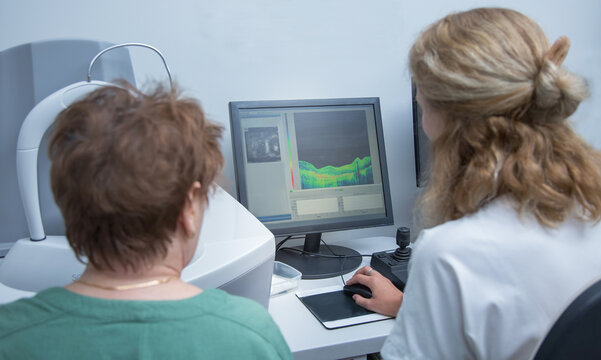 an elderly woman on a CT scanner does a retinal tomography in the oculist's office. brain, a female optometrist is sitting at a computer.
