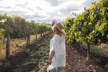 A beautiful middle aged woman looks at the expanses with a beatiful view of vineyards and mountain, senior dressed boho style and walks around a fiels with a vine