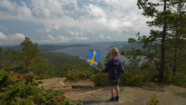 Boy waving Swedish flag atop Skuleberget  mountain overlooking breathtaking Nordic landscape. Static