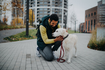 Happy young man stroking his dog outdoors in city park, during cold autumn day.