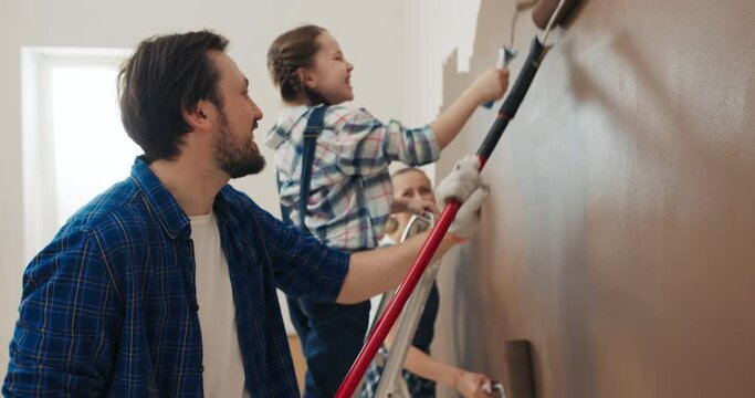Cheerful family has bought new house and is painting walls in brown. Man, wife and daughter with pigtails hold rollers in hands. The girl stands on the ladder and laughs loudly. They have fun.