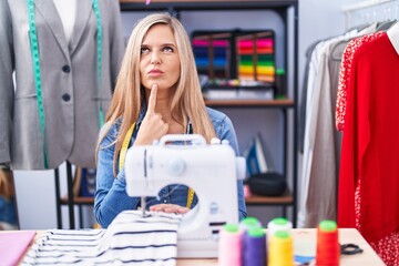 Blonde woman dressmaker designer using sew machine thinking concentrated about doubt with finger on chin and looking up wondering