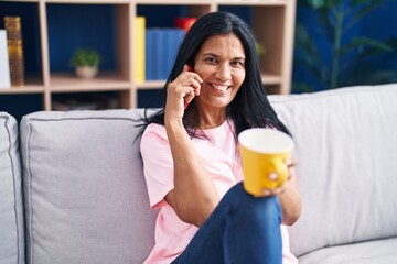 Middle age hispanic woman talking on smartphone drinking coffee at home
