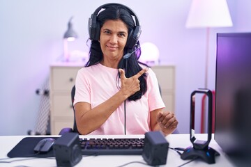 Mature hispanic woman playing video games at home cheerful with a smile of face pointing with hand and finger up to the side with happy and natural expression on face