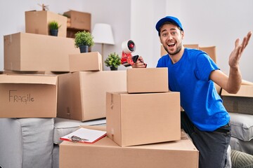 Hispanic man with beard working moving boxes celebrating victory with happy smile and winner expression with raised hands