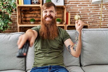Redhead man with long beard holding television remote control screaming proud, celebrating victory and success very excited with raised arm