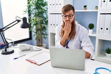 Redhead man with long beard wearing doctor uniform working using computer laptop covering mouth...