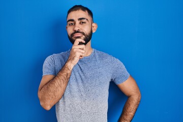 Middle east man with beard standing over blue background looking confident at the camera smiling with crossed arms and hand raised on chin. thinking positive.