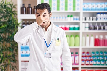 Handsome hispanic man working at pharmacy drugstore peeking in shock covering face and eyes with hand, looking through fingers with embarrassed expression.