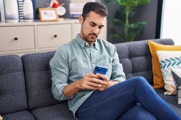 Young hispanic man using smartphone sitting on sofa at home