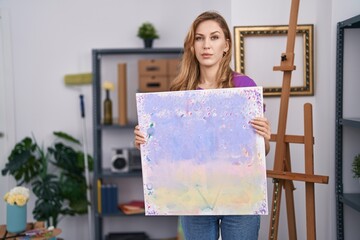 Beautiful blonde woman holding painting canvas skeptic and nervous, frowning upset because of...