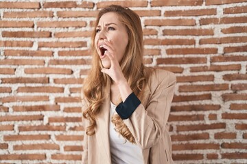 Beautiful blonde woman standing over bricks wall shouting and screaming loud to side with hand on mouth. communication concept.
