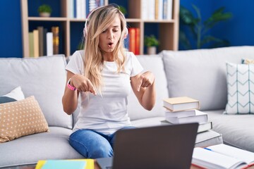 Obraz premium Young blonde woman studying using computer laptop at home pointing down with fingers showing advertisement, surprised face and open mouth
