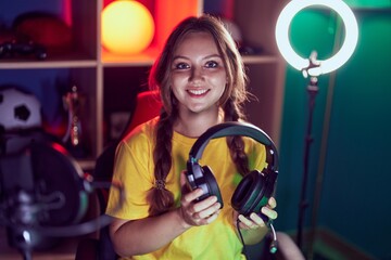 Young blonde woman streamer smiling confident holding headphones at gaming room