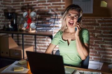 Young beautiful woman working at the office at night speaking on the phone sticking tongue out happy with funny expression. emotion concept.