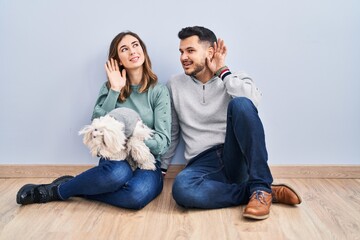 Young hispanic couple sitting on the floor with dog smiling with hand over ear listening an hearing...