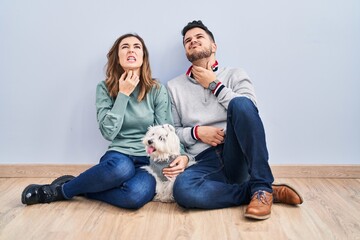 Young hispanic couple sitting on the floor with dog touching painful neck, sore throat for flu, clod and infection