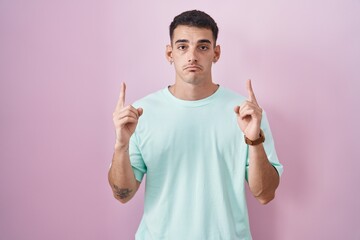 Handsome hispanic man standing over pink background pointing up looking sad and upset, indicating...