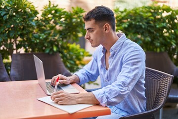 Young hispanic man using laptop writing on paper sitting on table at coffee shop terrace