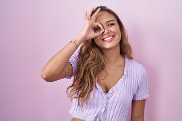Fototapeta premium Young hispanic woman standing over pink background doing ok gesture with hand smiling, eye looking through fingers with happy face.