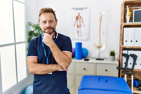 Middle age physiotherapist man working at pain recovery clinic looking confident at the camera with smile with crossed arms and hand raised on chin. thinking positive.