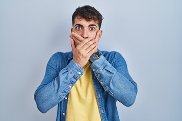 Young hispanic man standing over blue background shocked covering mouth with hands for mistake....