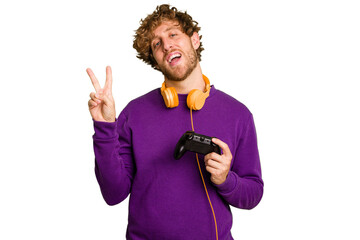 Young caucasian gamer man holding a game controller isolated on white background joyful and carefree showing a peace symbol with fingers. © Asier