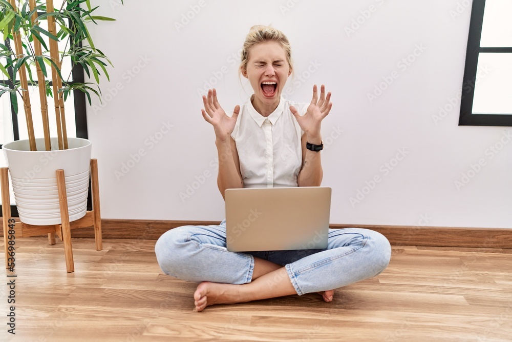 Canvas Prints Young blonde woman using computer laptop sitting on the floor at the living room celebrating mad and crazy for success with arms raised and closed eyes screaming excited. winner concept