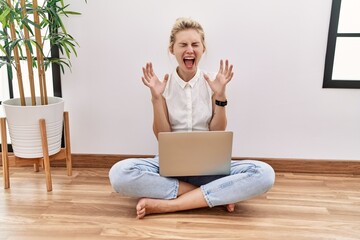 Young blonde woman using computer laptop sitting on the floor at the living room celebrating mad and crazy for success with arms raised and closed eyes screaming excited. winner concept