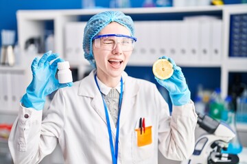 Beautiful woman working at scientist laboratory holding pills and lemon winking looking at the camera with sexy expression, cheerful and happy face.
