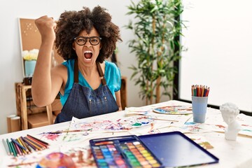 Fototapeta premium Beautiful african american woman with afro hair painting at art studio angry and mad raising fist frustrated and furious while shouting with anger. rage and aggressive concept.