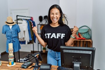 Young hispanic woman working as staff at retail boutique very happy and excited doing winner gesture with arms raised, smiling and screaming for success. celebration concept.