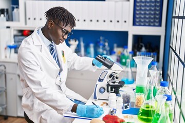 African american man scientist using microscope writing report at laboratory