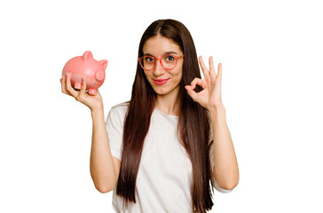 Young caucasian woman holding a piggy bank isolated cheerful and confident showing ok gesture.