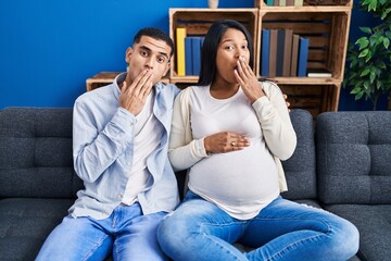 Young hispanic couple expecting a baby sitting on the sofa at home covering mouth with hand,...