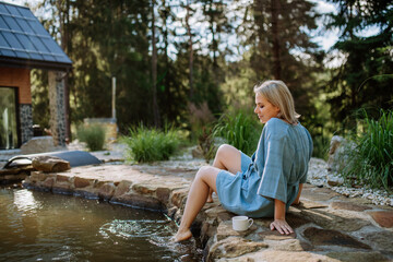Happy young woman sitting by pond near cottege and enjoying cup of morning coffee on summer vacation in mountains. High angle view.