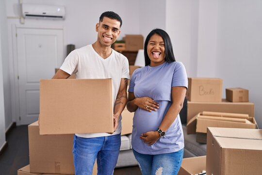 Young hispanic couple expecting a baby moving to a new home sticking tongue out happy with funny expression.