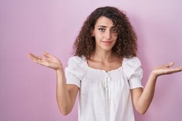 Hispanic woman with curly hair standing over pink background clueless and confused expression with...