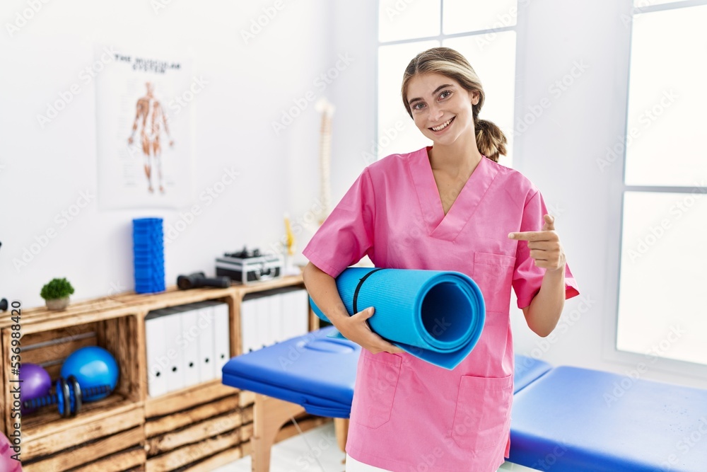 Poster Young physiotherapist woman working at pain recovery clinic holding yoga mat smiling happy pointing with hand and finger