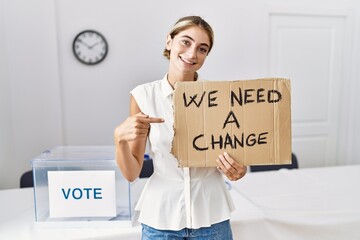 Young blonde woman at political election holding we need a change banner smiling happy pointing...