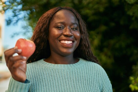 Young african woman smiling happy holding fresh red apple at the park