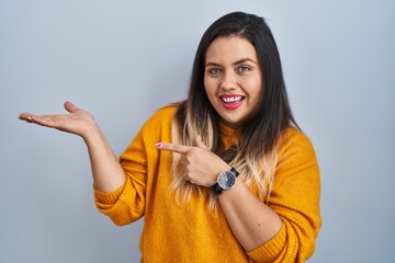 Young hispanic woman standing over isolated background amazed and smiling to the camera while presenting with hand and pointing with finger.