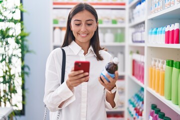 Young beautiful hispanic woman customer using smartphone holding medicine bottle at pharmacy
