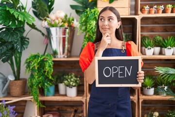 Young hispanic woman working at florist holding open sign serious face thinking about question with...
