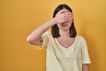 Hispanic girl wearing casual t shirt over yellow background covering eyes with hand, looking...