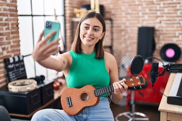 Young hispanic woman musician make selfie by the smartphone holding ukelele at music studio